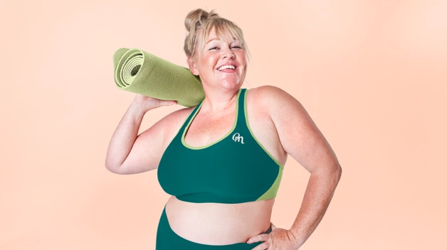 A women with obesity wearing workout clothes and holding a ypga mat in front of a pink background