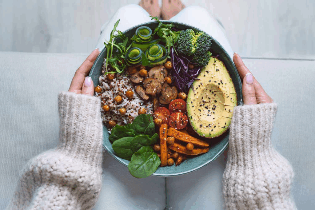 Woman in cozy sweater holding a colorful Buddha bowl with avocado, vegetables, chickpeas, and grains.