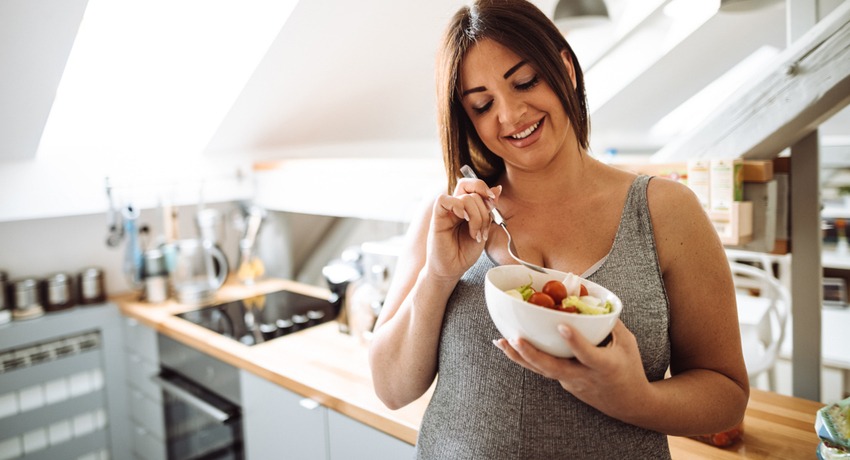 Smiling woman enjoying a bowl of fresh fruit salad in a bright, modern kitchen with a skylight.