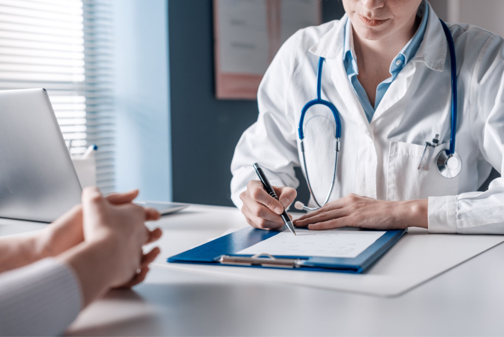 Doctor in a white coat with a stethoscope writing notes on a clipboard while consulting with a patient across the desk.