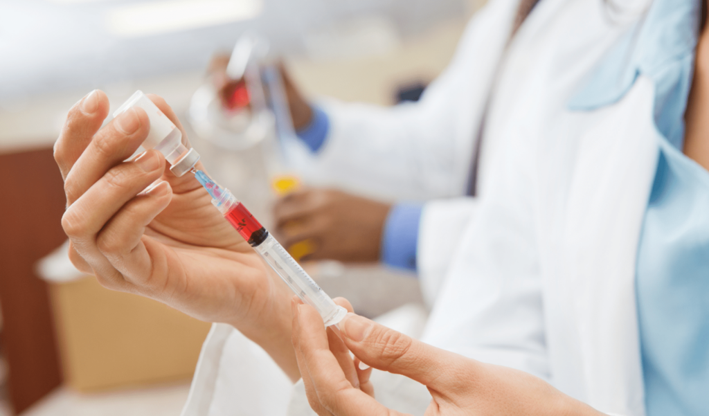 Healthcare worker in a white coat holding a blood sample tube or syringe with red liquid in a medical laboratory setting.