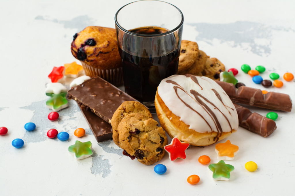 Assortment of sugary foods, including a glazed donut, muffin, cookies, chocolate bars, colorful candies, gummy stars, and a glass of soda on a light surface.