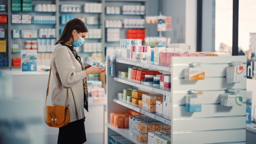 Masked woman reading a medication package label while browsing shelves stocked with boxed medicines at a retail pharmacy.