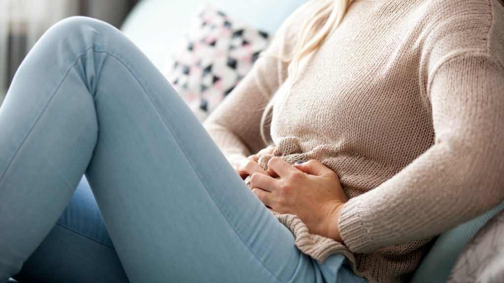Woman sitting on a couch, clutching her lower abdomen in discomfort, wearing a beige sweater and light blue jeans.