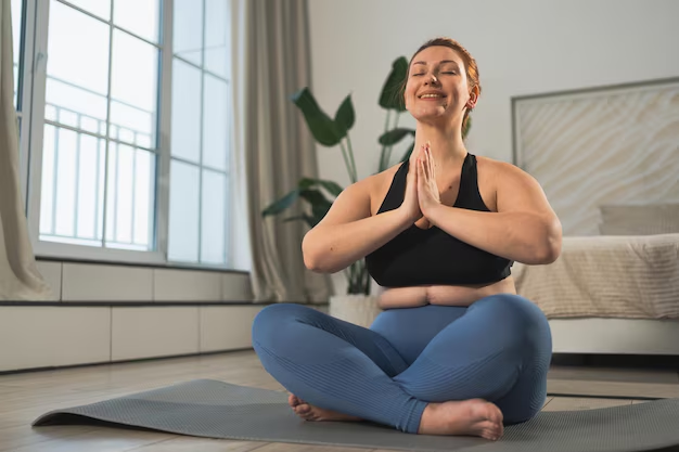Smiling woman sitting cross-legged on a yoga mat in her bedroom, hands in prayer pose, practicing mindful movement at home.