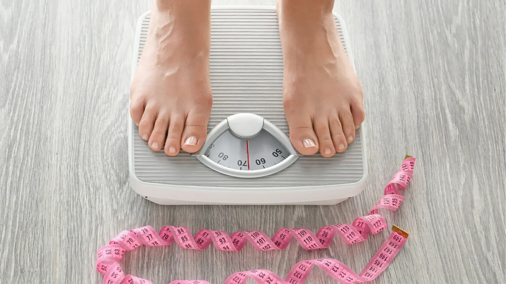 Person's bare feet on a white bathroom scale with pink measuring tape on a wooden floor, representing weight loss or health monitoring.